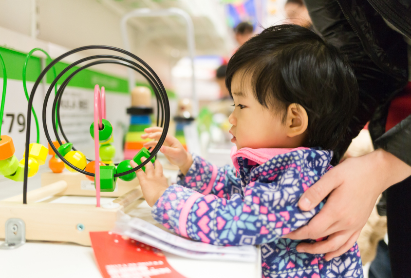 small toddler looks at colorful motor skills toy in store with mother