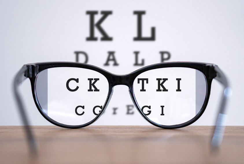 Photo: A pair of glasses sits in front of an eyesight testing poster with letters. The poster is magnified through the glasses lenses.