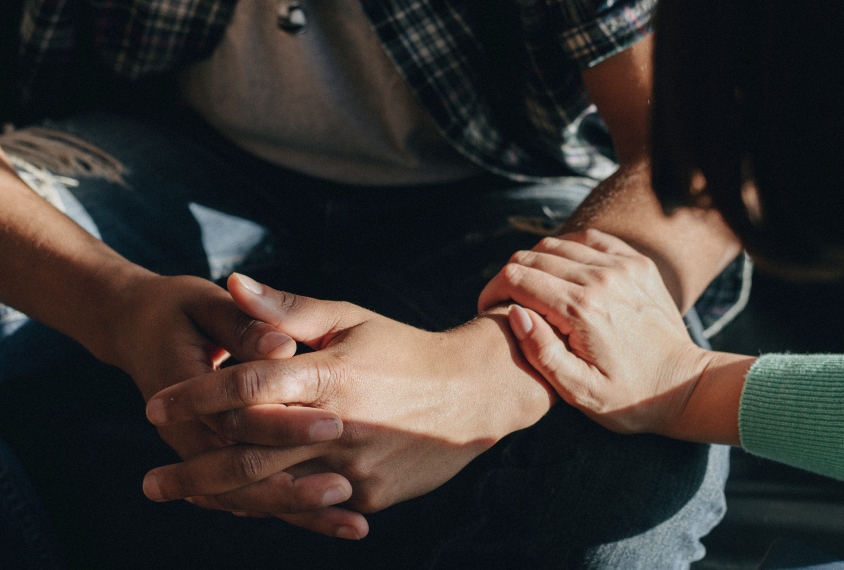 Close-up image of an arm reaching out to console someone who is sitting down with their hands folded in their lap.