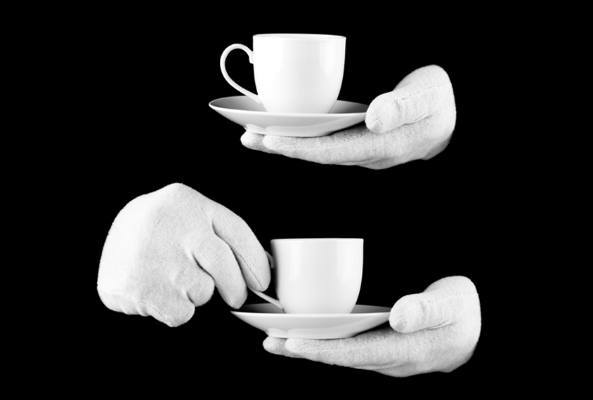 White gloved hands hold a white mug and white saucer against a black background.