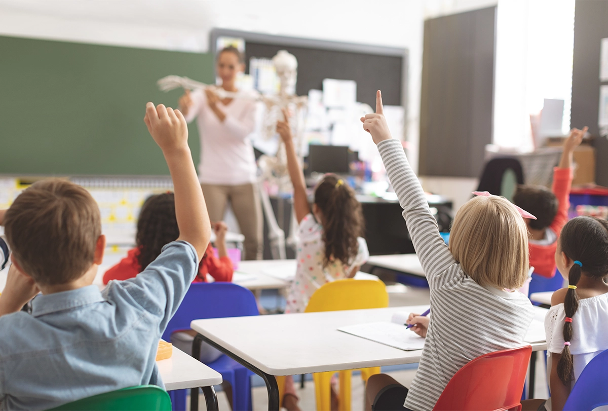 A classroom scene showing elementary kids raising hands at their desks.