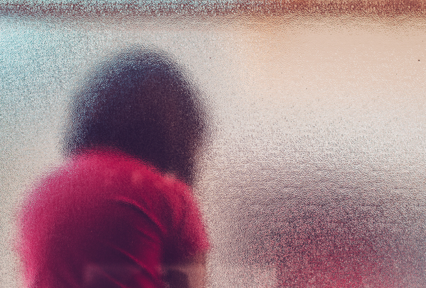 Child in sad posture, seen from through a textured glass window