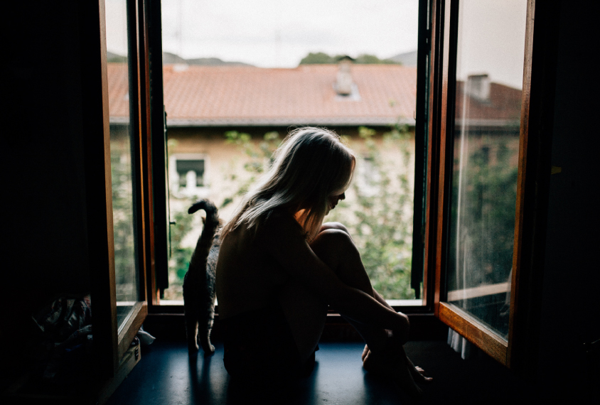 young woman or girl sits alone on a window sill, in a sad posture.