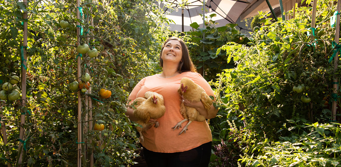 Holly Stessman standing in her garden looking up and holding her two chickens.