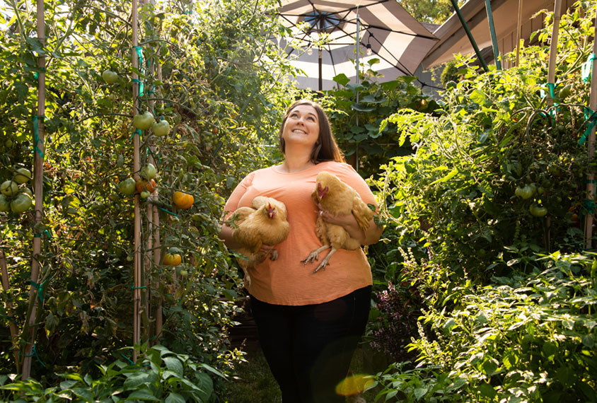 Holly Stessman standing in her garden looking up and holding her two chickens.