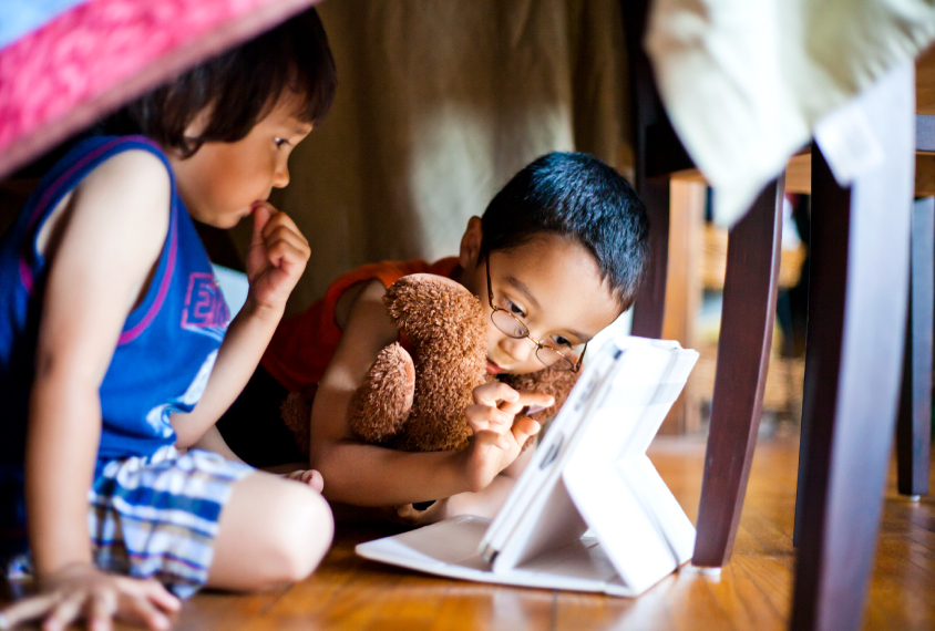 Two young children play with a tablet computer underneath a blanket fort