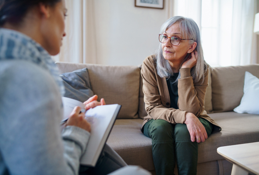 An older woman with gray hair sits on a couch and speaks with a clinician.