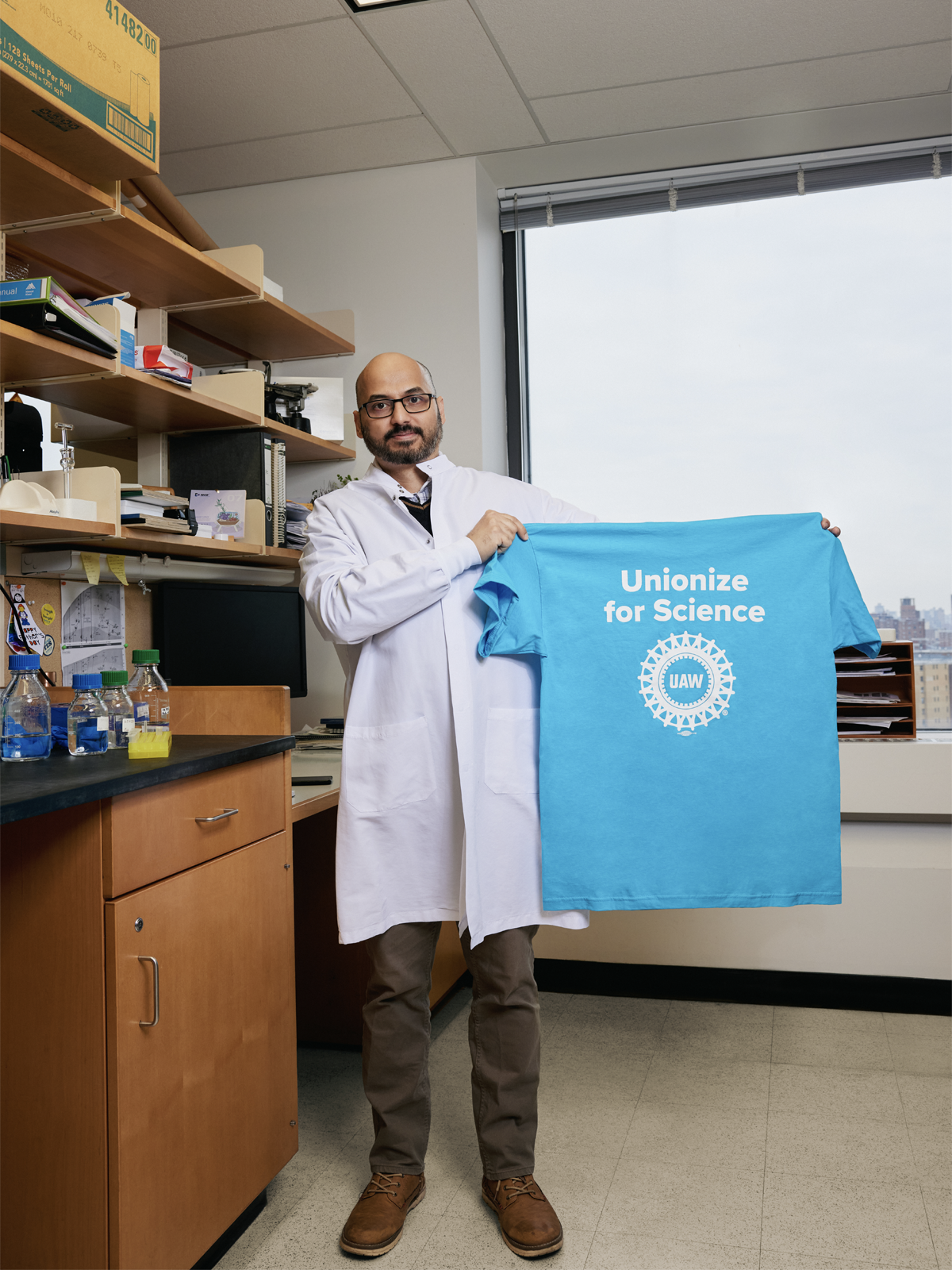 Balagopal Pai stands in a lab wearing a white coat and holding up a light blue t-shirt that says Unionize for Science above a United Auto Workers logo.