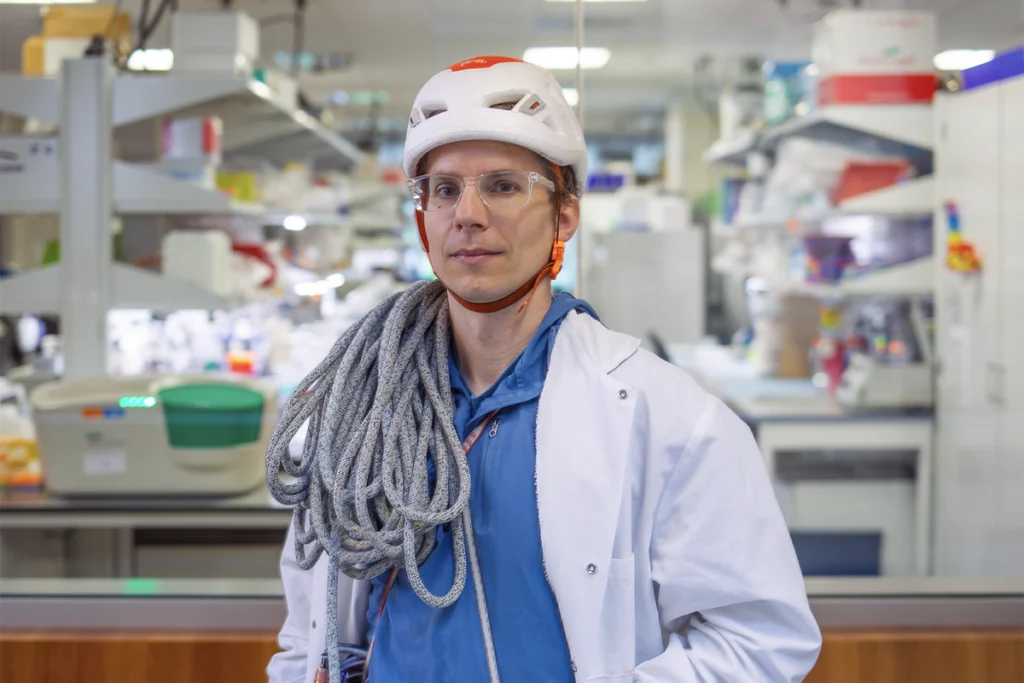 Portrait of Kaspar Podgorski standing in his lab wearing a helmet with a climbing rope over his shoulder.