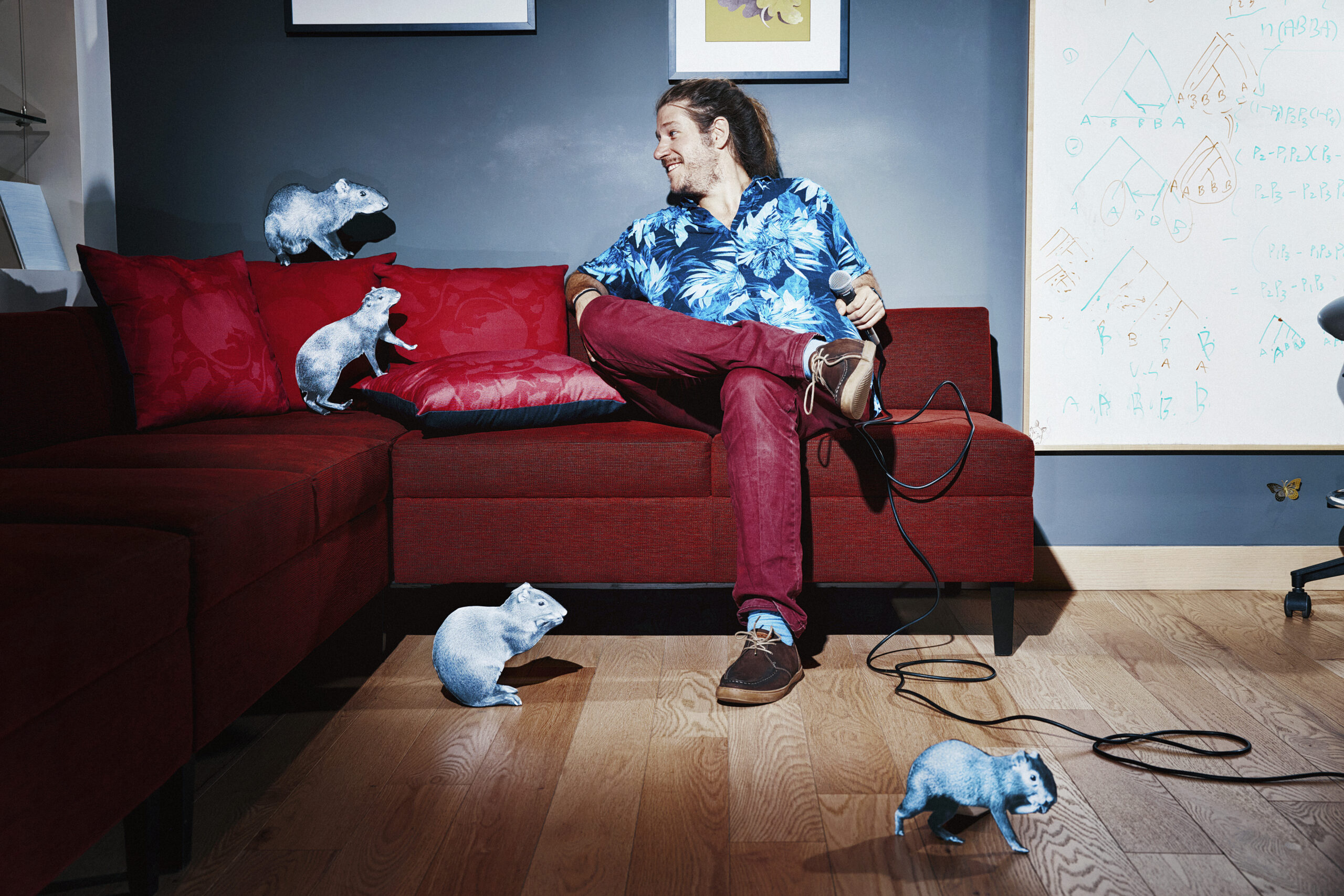 Neuroscientist Nacho Sanguinetti with a microphone on a red couch looking at black and white photo cutouts of realistic agoutis set up like an audience.