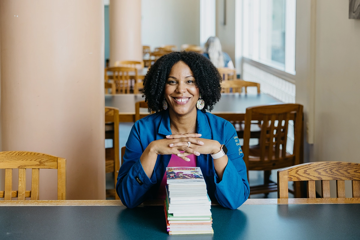 Photograph of Theanne Griffith sitting at a table with her hands interlocked over a stack of books, with one that she has published at the very top.