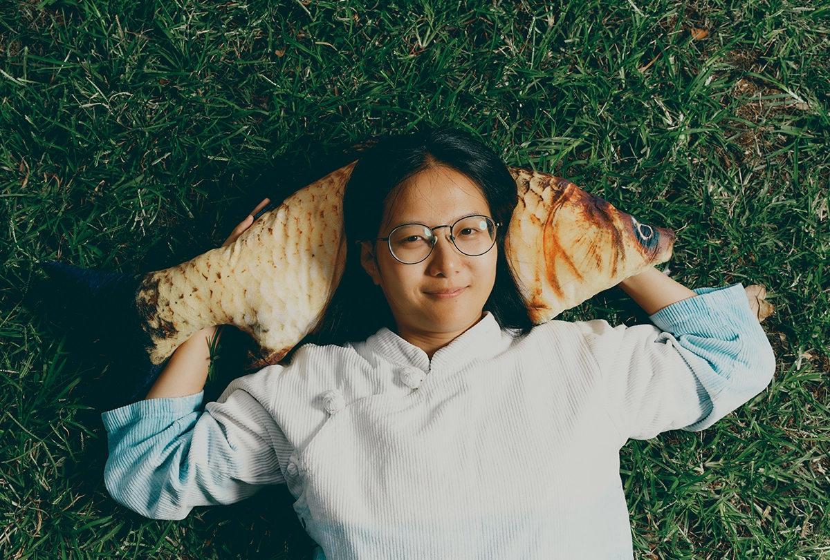 Young researcher laying in the grass holding a pillow shaped like a fish.