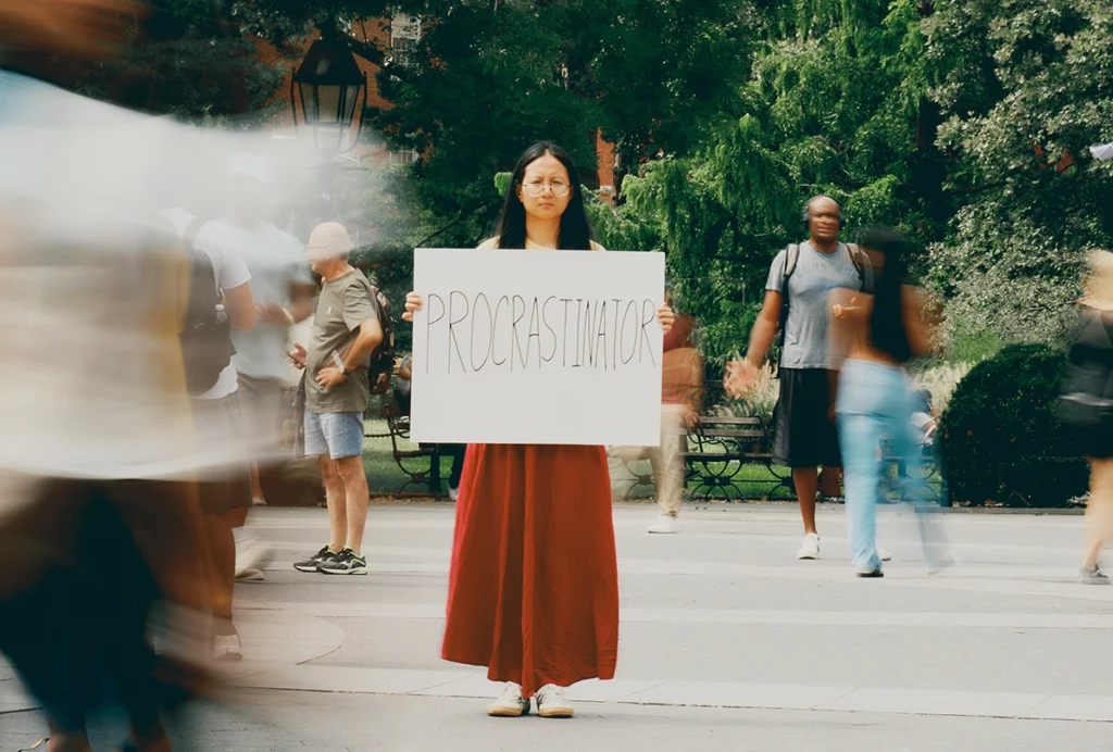 Photograph of Pei Yuan Zhang standing alone in a park holding up a white sign with "PROCRASTINATOR" written on it in black marker.