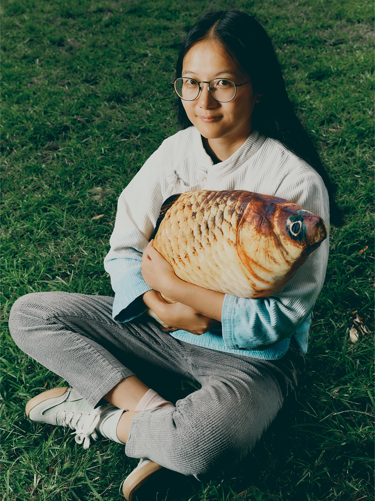 Young researcher sitting in the grass holding a pillow shaped like a fish.