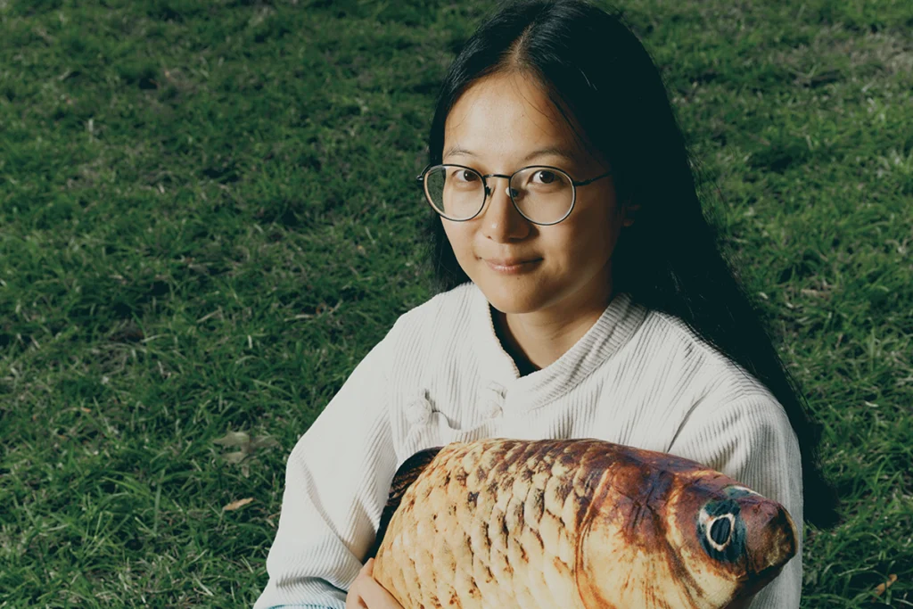 Young researcher sitting in the grass holding a pillow shaped like a fish.