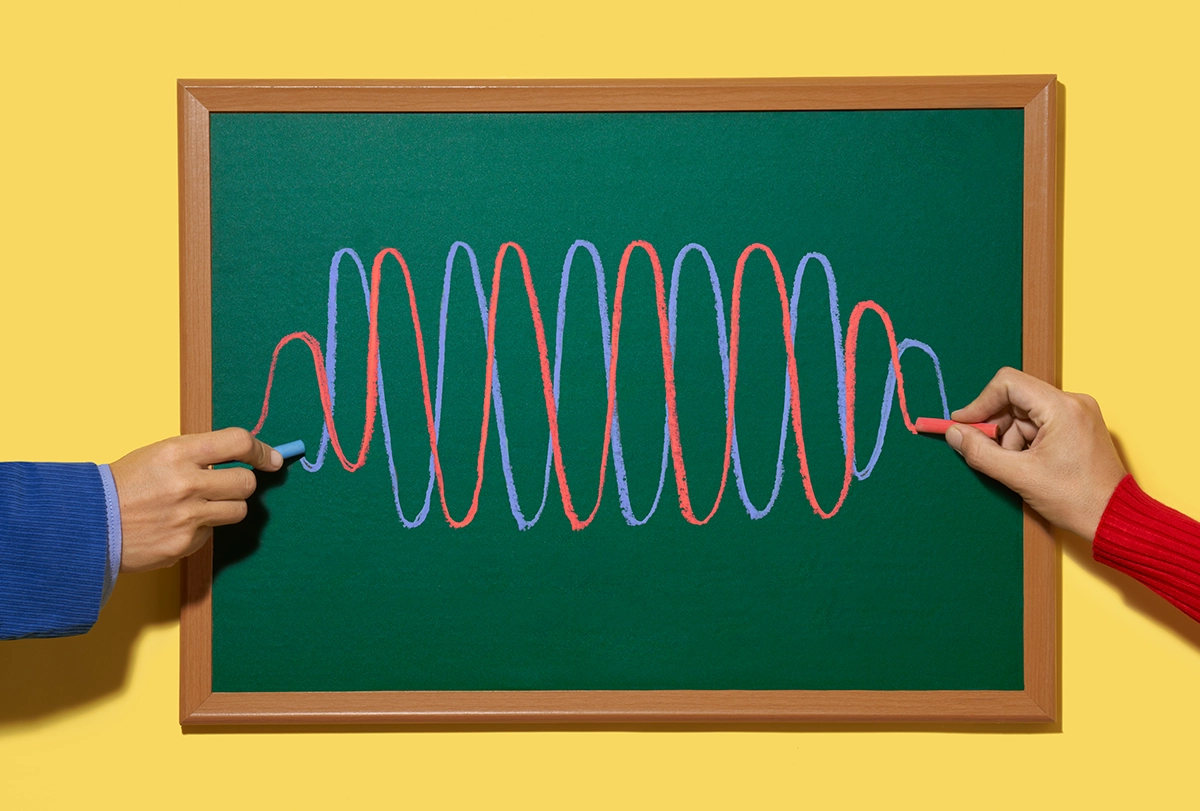 Photograph of two hands drawing overlapping red and blue waveforms on a chalkboard.