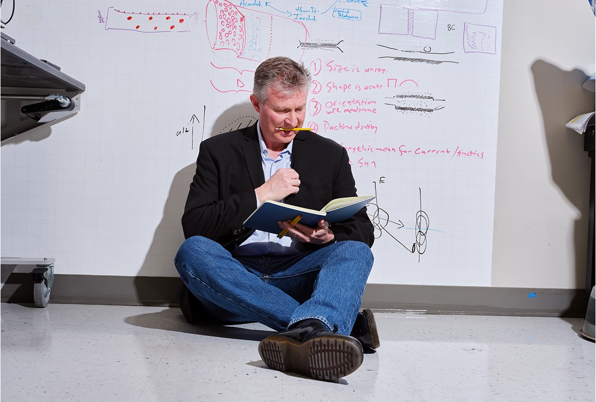 Bryan W Jones sits with his back against a large sheet of gridded paper, holding a notebook in his hand and a pencil in his mouth.