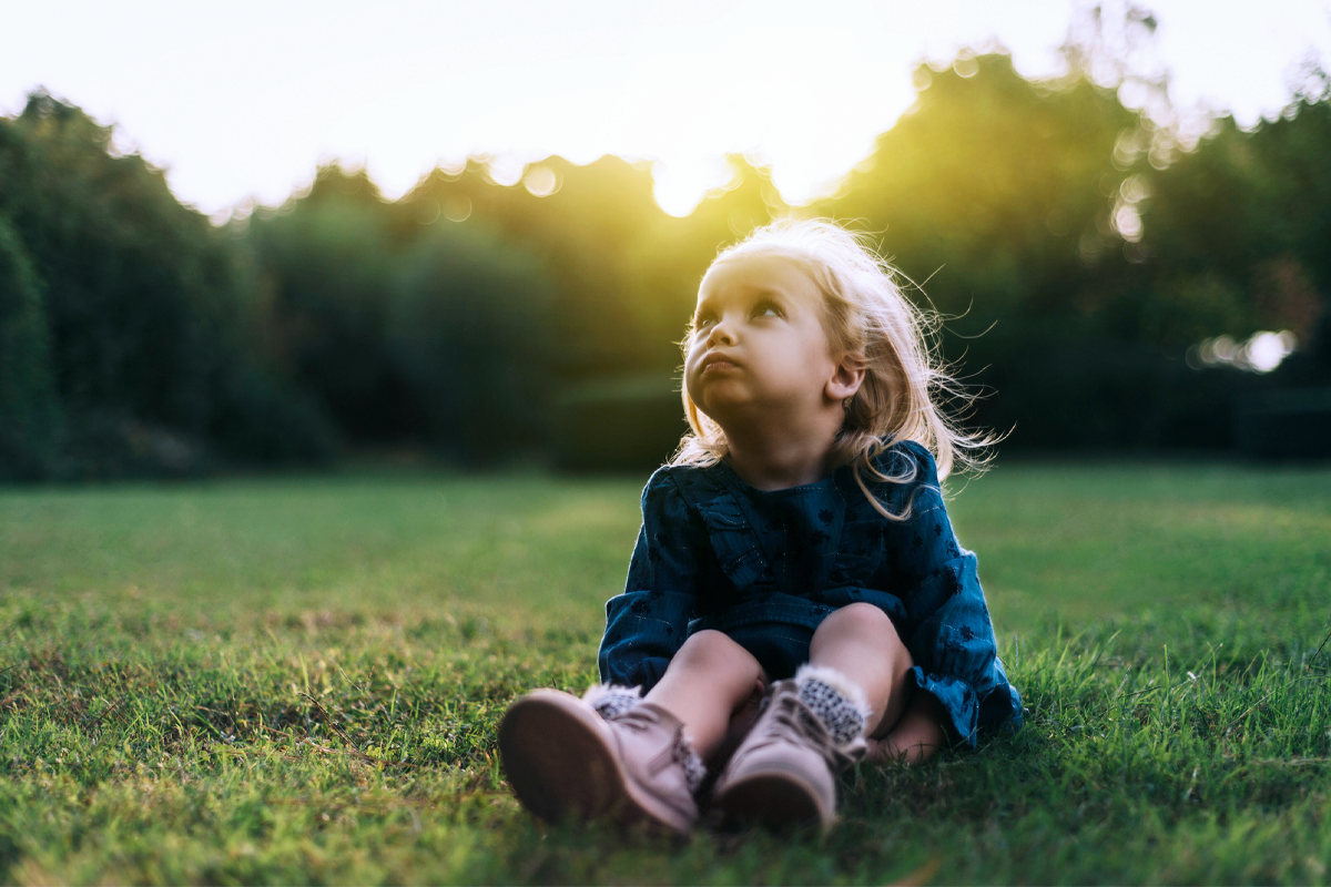 A sad-looking toddler sits in a field and looks up at the sky.