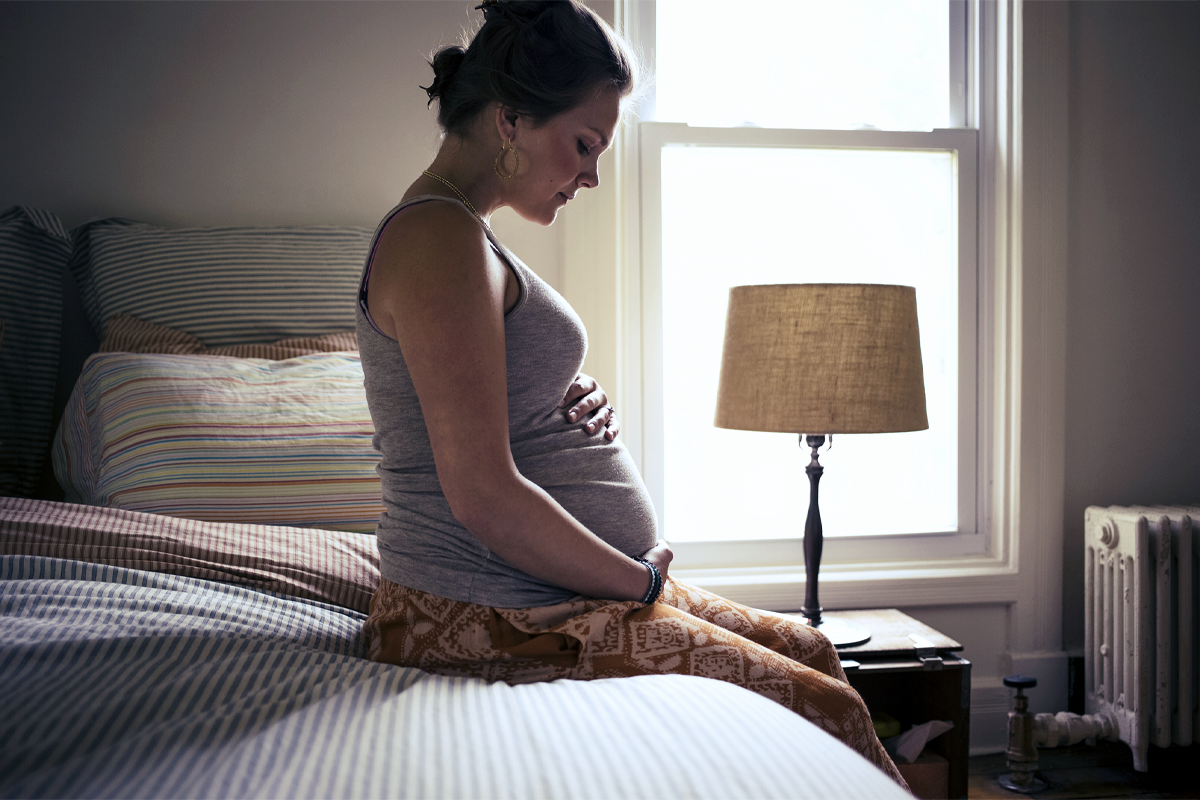 A pregnant woman seen in profile, sitting on the edge of a bed.